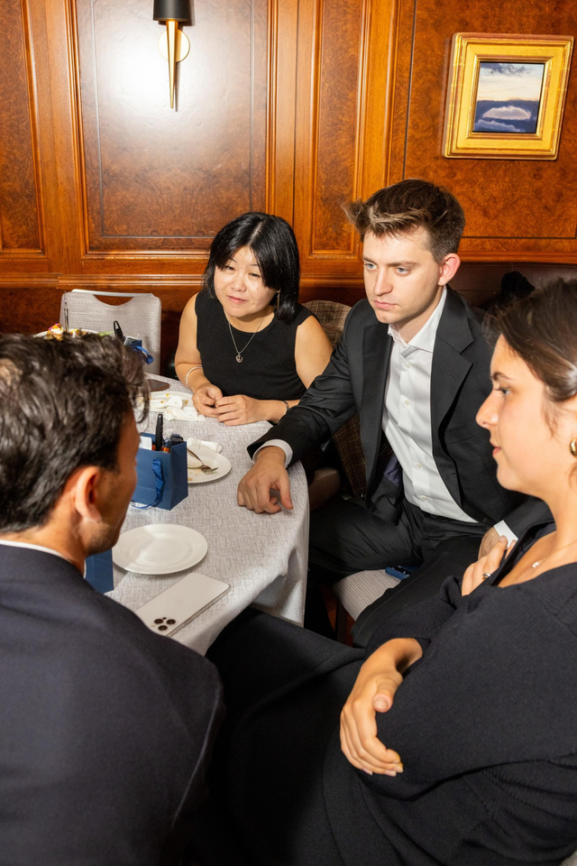 Four people in formal attire sit around a table in a wood-paneled room, engaged in a serious conversation.